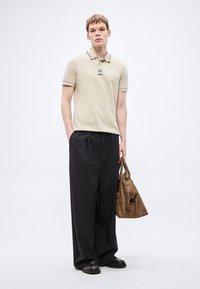 Κρεμ πόλο μπλουζάκι με μαύρες λεπτομέρειες, συνδυασμένο με άνετο μαύρο παντελόνι. Το μοντέλο κρατάει καφέ δερμάτινη τσάντα, στεκόμενο μπροστά σε ανοιχτόφωρο φόντο.