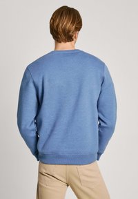 Sudadera azul con cuello redondo y puños acanalados. Textura suave con un ajuste relajado, combinada con pantalones beige. Vista trasera solamente.