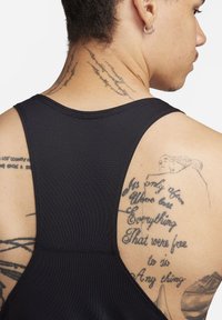 Schwarzes Sporttanktop mit Racerback-Design, das sichtbare Nähte sowie Tätowierungen auf dem Rücken und der Schulter zeigt. Glatte Textur.