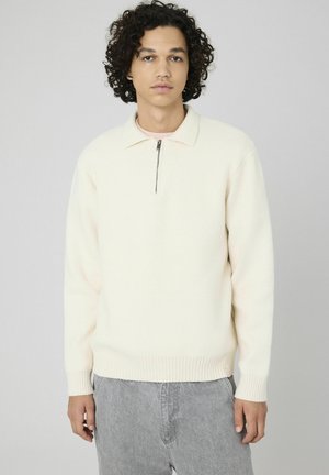 Pull en tricot beige clair avec une fermeture éclair au col, des poignets et un ourlet côtelés, présentant une texture douce et une coupe décontractée. Assorti avec un pantalon gris.