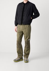 Chaqueta bomber negra con cuello acanalado, pantalones verdes olivo con parches oscuros en las rodillas y zapatillas grises con suelas verde lima.