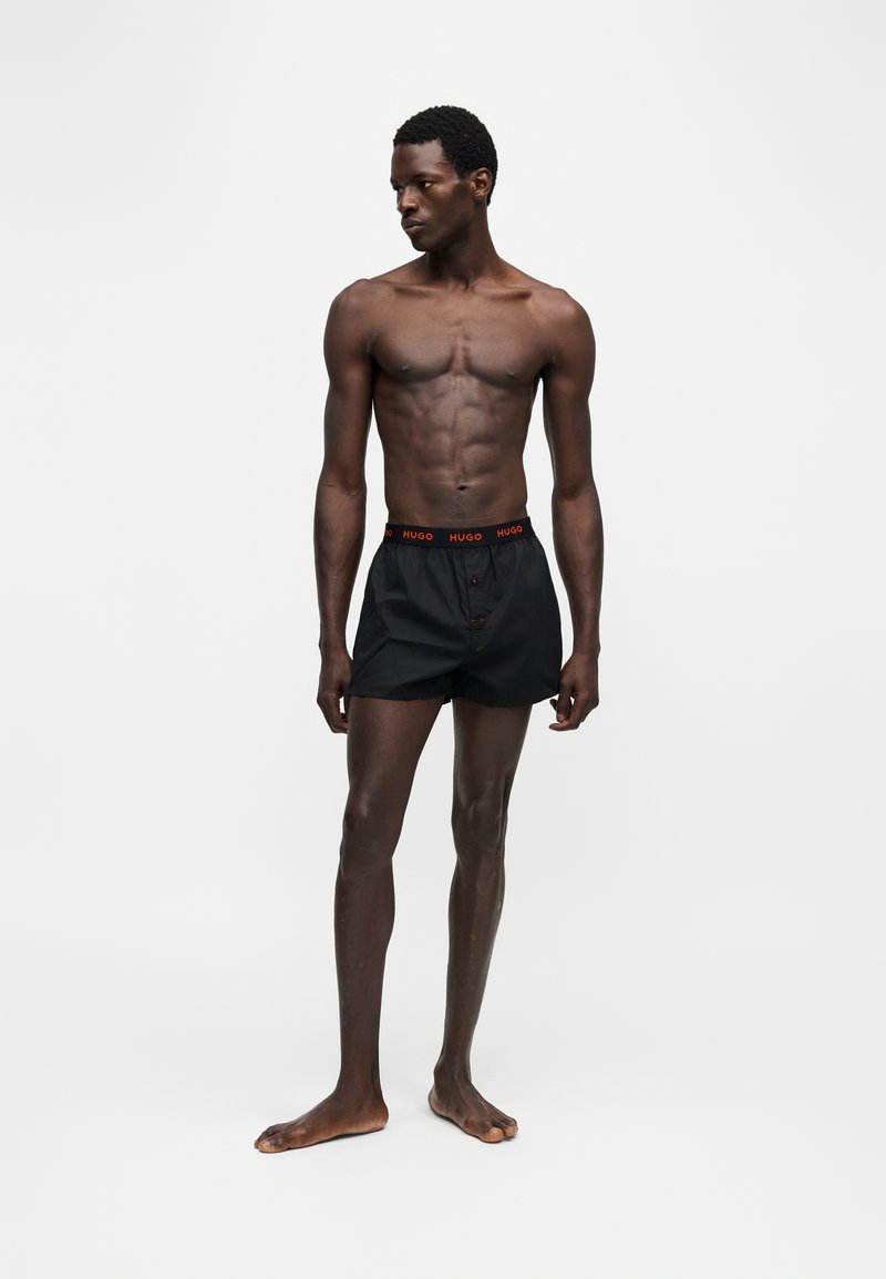 Homme torse nu debout, portant un boxer noir avec un logo rouge "HUGO" à la ceinture, regardant vers sa gauche sur fond uni.