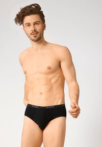 Schwarze Sloggi Slips mit einem grauen Logo-Bund, im klassischen Slip-Design und aus glattem Stoff. Das Modell steht in Pose.
