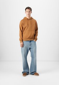 Sweatshirt à capuche marron avec une poche avant, associé à un jean flare bleu clair et des chaussures beiges à enfiler, présenté sur un fond blanc.