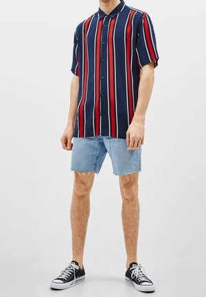 Uomo che indossa una camicia blu navy a maniche corte con bottoni, a righe verticali rosse e bianche, pantaloncini di jeans chiari e scarpe sportive nere basse.