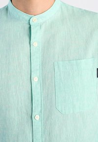 Camicia in lino di un azzurro chiaro con colletto a banda, bottoni frontali e tasca sul lato sinistro del petto. Tessuto morbido e design casual.