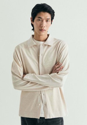 Joven con cabello oscuro, de pie con los brazos cruzados, vistiendo una camisa beige con botones sobre una camiseta blanca, frente a un fondo claro y liso.