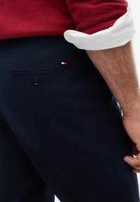 Homme portant un pantalon bleu marine foncé avec un petit logo rouge, blanc et bleu près d'une poche arrière et un pull rouge avec les manches retroussées.
