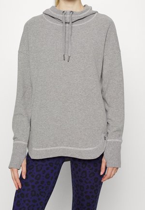 Vrouw die een lichtgrijze pullover met capuchon en trekkoorden draagt, en donkerblauwe legging met een zwart luipaardprintpatroon.