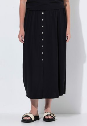 Person iført en sort midi-nederdel med knapper foran og creme- og sorte slip-in sandaler, der står op ad en hvid væg.