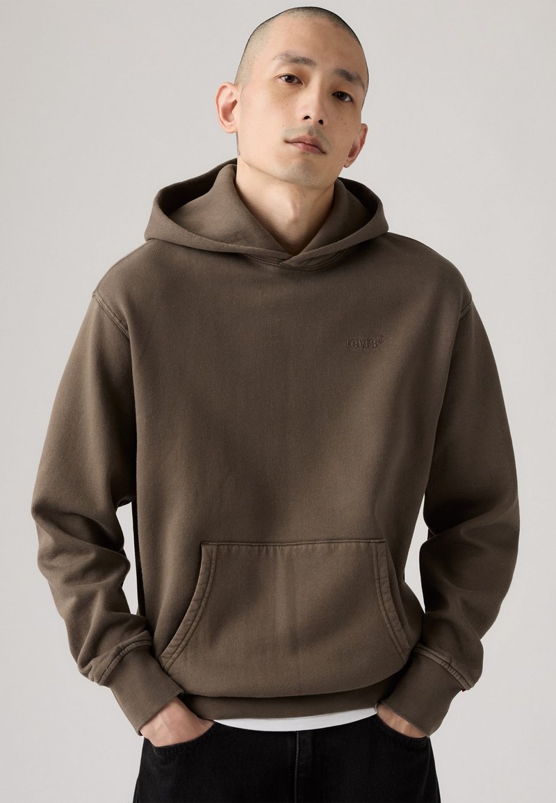 Sudadera con capucha marrón hecha de una suave mezcla de algodón. Presenta un bolsillo tipo canguro y un logo bordado en el pecho. Textura suave.