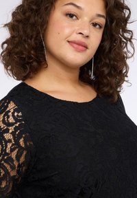 Schwarzes Spitzenoberteil mit floralem Muster, das einen runden Ausschnitt und durchsichtige lange Ärmel aufweist. Das Modell zeigt texturierten Stoff und dezente Details.
