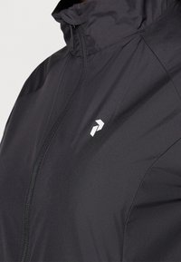 Veste coupe-vent légère noire avec col montant, fermeture éclair et logo blanc sur la poitrine gauche. Texture lisse, coupe ajustée.