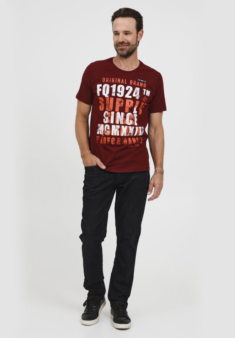 Camiseta de algodón burdeos con un gráfico de texto en blanco y naranja en bold, combinada con pantalones de mezclilla negros y zapatillas negras. Diseño simple y casual.