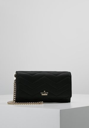 Sort quiltet pung med gyldent Kate Spade-logo og kæderem, der hviler på en hvid podium mod en grå baggrund.