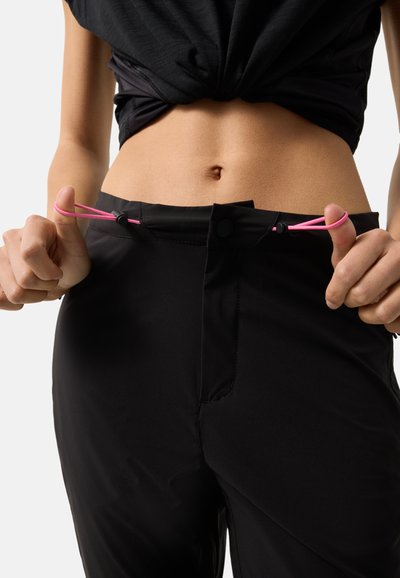 Nahaufnahme einer Person, die den Bund einer schwarzen Hose mit rosa elastischen Kordeln anpasst, dabei trägt sie ein schwarzes Oberteil, das hochgezogen ist und den Bauch freilegt.