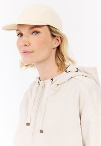Beige baseballpet met een gebogen rand, voorzien van een gladde stoftextuur. Gemaakt met een lichtgekleurde, rits-hakende hoodie met een trekkoord.