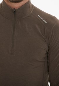 Bruine quarter-zip pullover met hoge kraag, gemaakt van zacht, rekbaar materiaal. Voorzien van platlock naden en het witte "ENDURANCE" logo.
