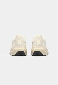 Beige och grå sneaker med en texturerad ovandel och mesh-accenter, med en dämpad sula och svart gummisula, sedd bakifrån.