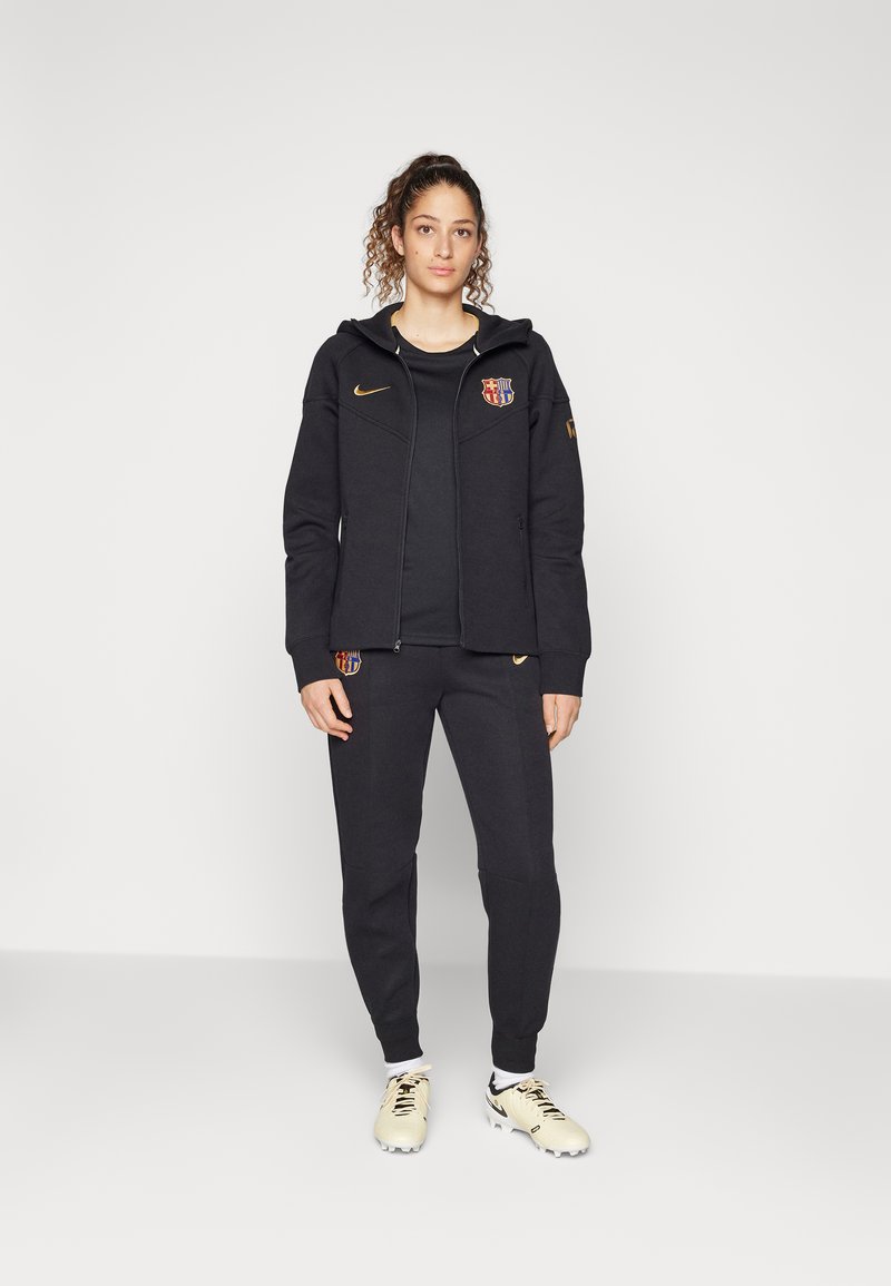 Felpa nera con zip e cappuccio, caratterizzata da un logo sul petto, abbinata a pantaloni neri coordinati e scarpe sportive gialle. Tessuto strutturato.