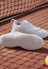 Scarpe sportive bianche con suole testurizzate su un campo da tennis in terra rossa vicino a una rete e una pallina da tennis gialla parzialmente visibile.