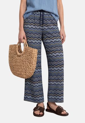 Kvinde iført blå zigzag-mønstrede bukser, blå top, brune sandaler, holder en vævet stråtaske ved sin side.
