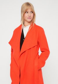 Mulher loira vestindo um casaco envolvente laranja brilhante com grandes lapelas sobre uma blusa preta e branca, olhando para a sua esquerda com as mãos nos bolsos.