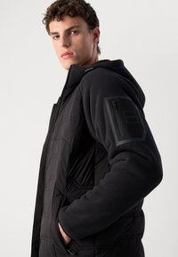 Zwarte gewatteerde jas met een fleece mouw, voorzien van een ritszak op de arm en textuurpatronen op het lichaam.