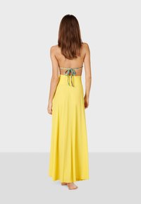 Vestido amarillo maxi con un diseño fluido, lazos en la parte trasera y cintura alta. Tejido suave, minimalismo en los adornos y sin mangas.