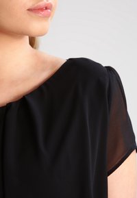 Černá blůza s krátkými, průhlednými rukávy a kulatým výstřihem. Tkanina vypadá hladce, s jemným skládáním u ramene.