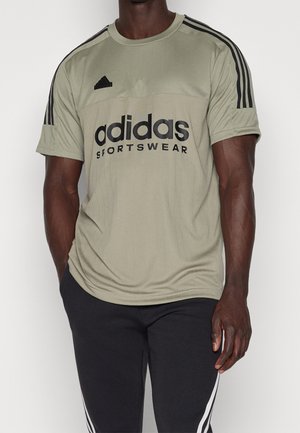 Mann trägt ein beiges Adidas-Sportshirt mit schwarzen Streifen auf den Schultern und eine schwarze Adidas-Hose mit weißen Streifen.