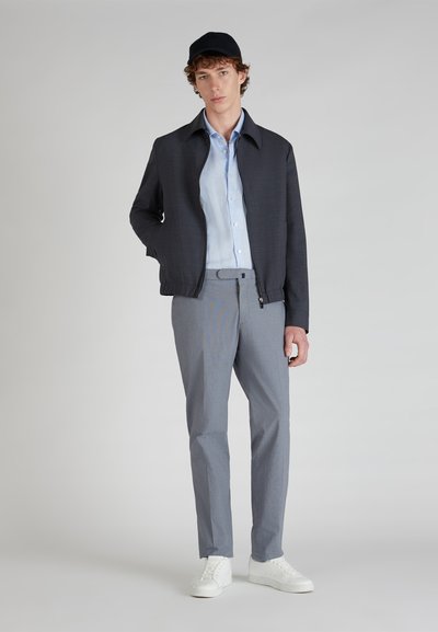 Giacca scura da uomo, camicia azzurro chiaro, pantaloni grigi a fantasia e sneakers bianche. Tessuto morbido, chiusura con zip e colletto strutturato. Cappellino nero.