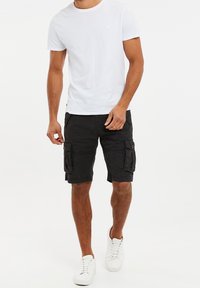 Un t-shirt blanc en coton pour homme associé à un short cargo noir avec deux poches latérales et des accents de boutons. Complété par des baskets blanches.