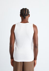 Weißes, geripptes Tanktop mit tailliertem Design, das eine glatte Textur und einen Standard-Rückenaufschnitt zeigt, kombiniert mit gemusterten braunen Karoshorts.