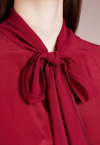 Primo piano di una blusa rossa con un fiocco legato al colletto, indossata da una persona con il collo visibile e lunghi capelli castani.