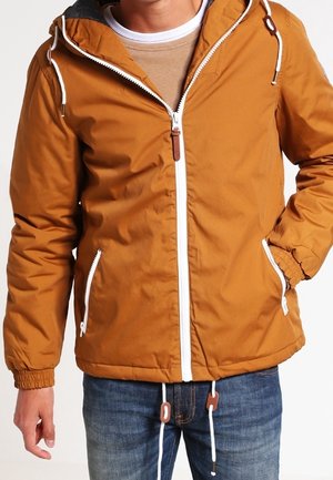 Uomo che indossa una giacca marrone con cappuccio e cerniera, con coulisse bianche, abbinata a jeans blu e un maglione beige sotto.