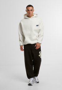 Hanorac alb cu buzunar și logo albastru, asociat cu pantaloni de fleece maro închis cu un design crem. Pantofii sport completează ținuta.