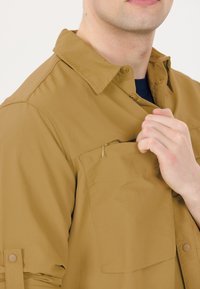 Tan overhemd met kraag, voorzien van een ritssluiting op de borstzak, een zachte textuur en opgerolde mouwen die zijn vastgezet met knooptabbladen.