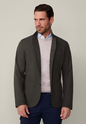Mørkegrøn blazer af glat stof, med en lille krave, to forsidelommer og en enkelt knaplukning, lagd over en lys sweater.