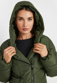 Zielona pikowana kurtka z zamkiem z przodu i regulowanym kapturem. Posiada elastyczne mankiety oraz gładką teksturę z pikowanym wykończeniem.