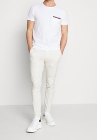 Biała koszulka z krótkim rękawem i kieszonką na piersi, ozdobiona czerwonymi i granatowymi paskami, w zestawie z jasnobeżowymi spodniami typu slim fit i białymi trampkami.