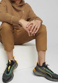 Person, der braune Hose und Kapuzenpulli trägt, sitzt mit gefalteten Händen und trägt schwarze, grüne und gelbe Sportschuhe auf einem weißen Block.