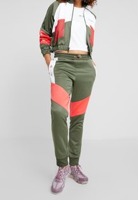 Frau in einem grün und korallenfarbigen Color-Block-Trainingsanzug mit einem weißen Cropped-Top und rosa transparenten Sneakers, die mit Händen in den Taschen steht.