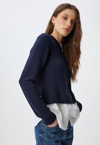 Pull en mailles de couleur marine avec un col rond, des manches longues et des poignets côtelés, porté par-dessus une chemise blanche et associé à un jean bleu.