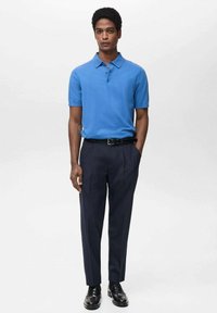 Cămașă polo albastră dintr-un material ușor, cu mâneci scurte, guler cu două nasturi; asortată cu pantaloni navy eleganți și pantofi negri.