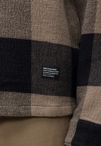 Gros plan sur un tissu à carreaux marron et noir avec une petite étiquette noire portant la mention "Marc O'Polo Denim from Stockholm, Sweden, Authentic Scandinavian."