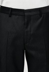Pantalon noir sur mesure avec un motif paisley subtil et des passants de ceinture, porté avec un top blanc rentré.