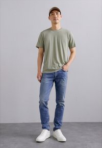 T-shirt en coton vert, jeans en denim bleu clair et baskets blanches. Un mannequin se tient droit contre un fond gris neutre.