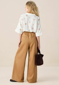 Witte blouse met schelpenpatronen en gouden accenten, gecombineerd met beige wijde broek. Houdt een kleine bruine tas vast met cirkelvormige uitsparingen.