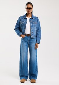 Jeans jakna s ovratnikom, svijetloplave boje, u kombinaciji s širokim plavim trapericama. Smeđi pojas i smeđe čizme. Žena nosi tamne sunčane naočale.